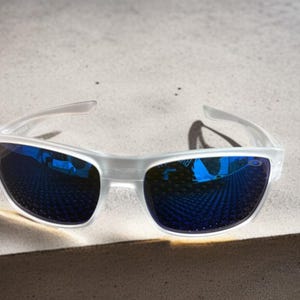 May include: Clear-framed sunglasses with blue mirrored lenses. The sunglasses are positioned on a light-colored surface, casting a shadow. The lenses reflect the surrounding environment, creating a vibrant blue hue.