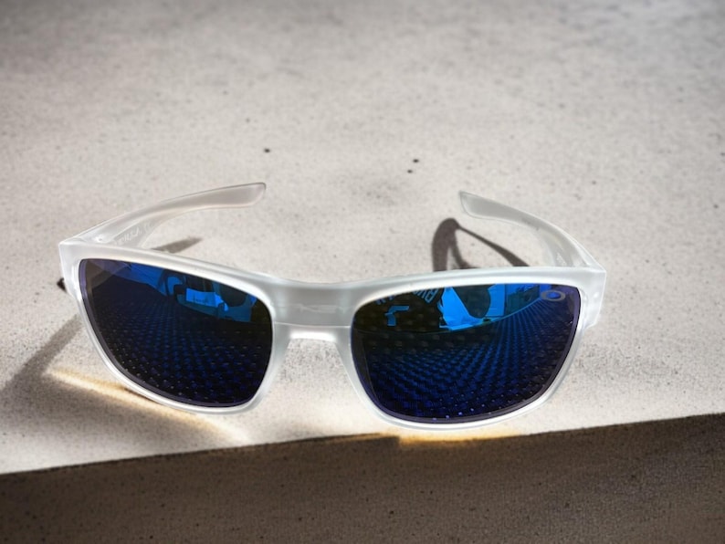 May include: Clear-framed sunglasses with blue mirrored lenses. The sunglasses are positioned on a light-colored surface, casting a shadow. The lenses reflect the surrounding environment, creating a vibrant blue hue.