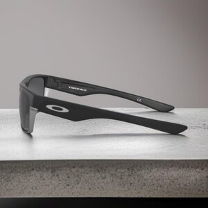 May include: Black Oakley sunglasses with the word "TWINFACE" printed on the frame. The sunglasses have a rectangular shape and a silver accent on the side. The Oakley logo is visible on the arm.