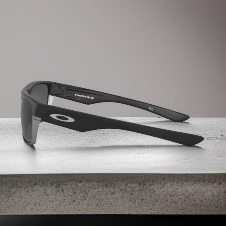May include: Black Oakley sunglasses with the word "TWINFACE" printed on the frame. The sunglasses have a rectangular shape and a silver accent on the side. The Oakley logo is visible on the arm.
