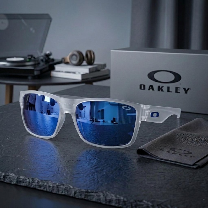 May include: Clear-framed sunglasses with blue lenses, resting on a dark surface. The Oakley logo is visible on the frame and a gray box. A cleaning cloth with the Oakley logo is also present.