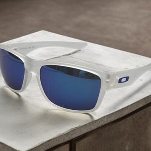 May include: White-framed sunglasses with blue mirrored lenses. The sunglasses have a rectangular shape and a small blue logo on the temples. The glasses are resting on a white surface.