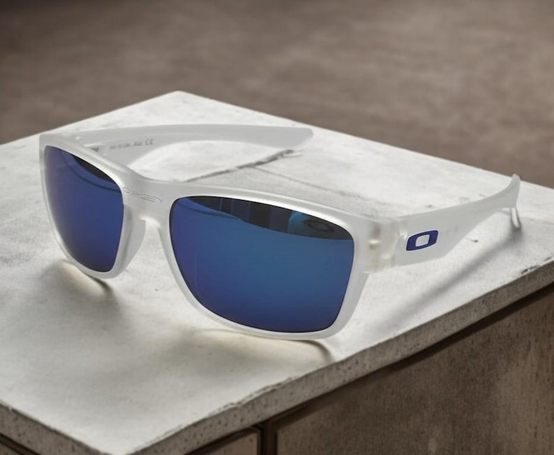 May include: White-framed sunglasses with blue mirrored lenses. The sunglasses have a rectangular shape and a small blue logo on the temples. The glasses are resting on a white surface.