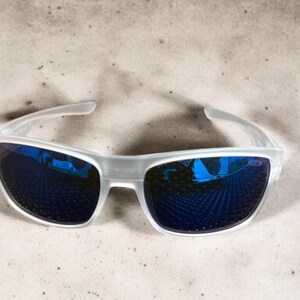 May include: White-framed sunglasses with blue mirrored lenses. The sunglasses have a modern, sporty design. The lenses reflect a blue hue, and the frame is a translucent white. The sunglasses are suitable for outdoor activities.