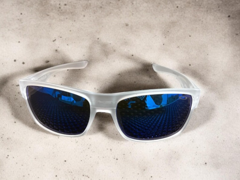 May include: White-framed sunglasses with blue mirrored lenses. The sunglasses have a modern, sporty design. The lenses reflect a blue hue, and the frame is a translucent white. The sunglasses are suitable for outdoor activities.