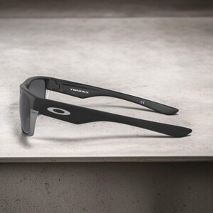 May include: Black sunglasses with gray lenses. The sunglasses have a rectangular frame and the text "TWINFACE" is visible on the temple. The Oakley logo is on the side of the frame.