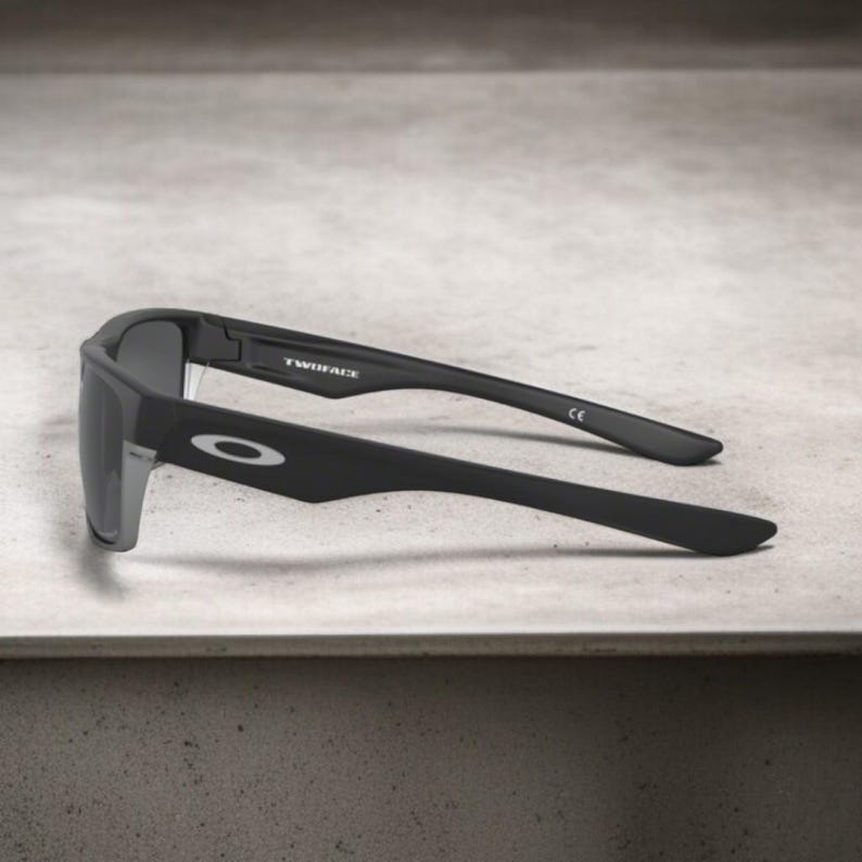 May include: Black sunglasses with gray lenses. The sunglasses have a rectangular frame and the text "TWINFACE" is visible on the temple. The Oakley logo is on the side of the frame.