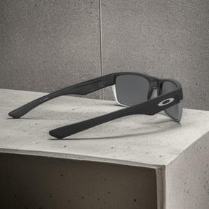 May include: Black sunglasses with gray lenses, resting on a concrete surface. The sunglasses have a rectangular frame and the Oakley logo is visible on the temple. The arms of the sunglasses are angled downward.