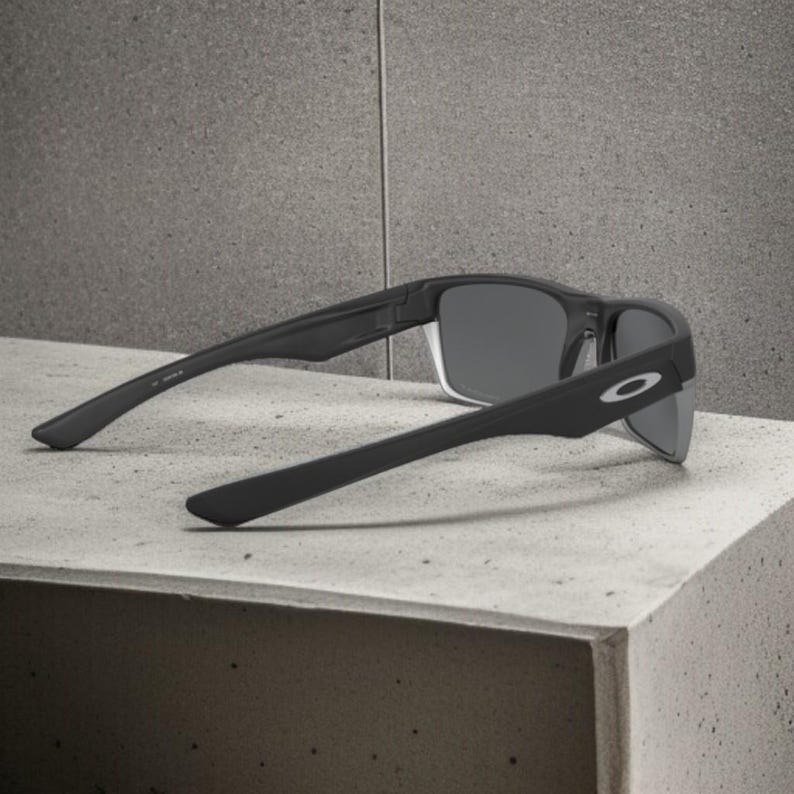 May include: Black sunglasses with gray lenses, resting on a concrete surface. The sunglasses have a rectangular frame and the Oakley logo is visible on the temple. The arms of the sunglasses are angled downward.