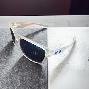 May include: Clear-framed sunglasses with blue lenses. The sunglasses have a sporty design with a curved frame and a small blue logo on the temple. The lenses reflect the surrounding environment.