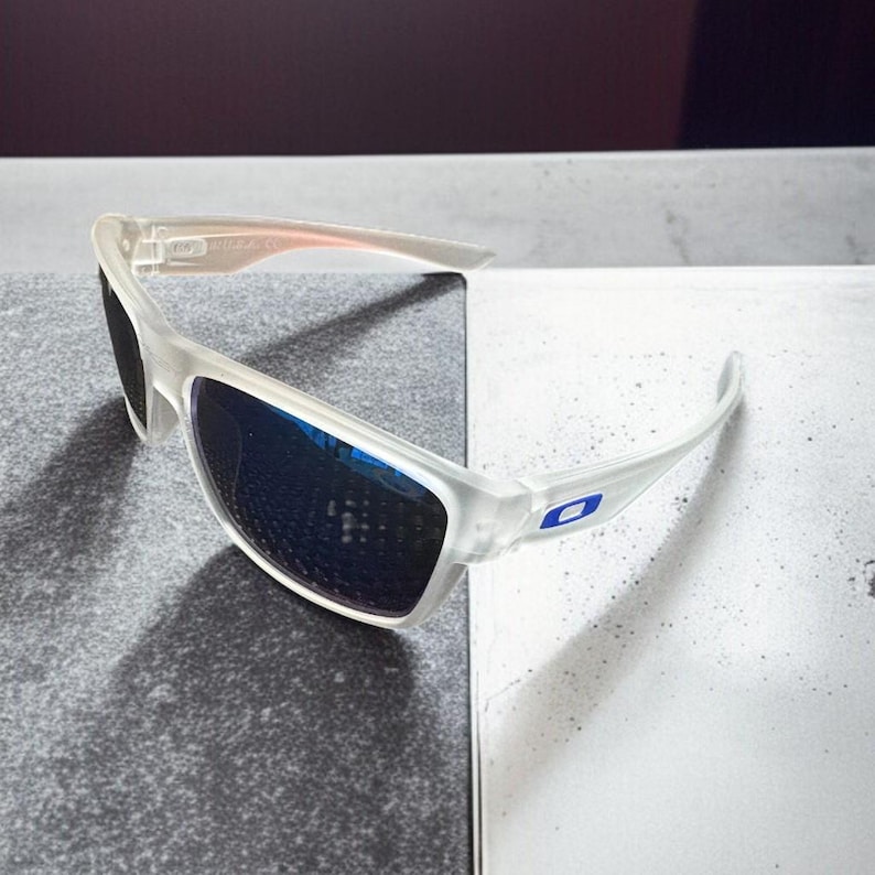 May include: Clear-framed sunglasses with blue lenses. The sunglasses have a sporty design with a curved frame and a small blue logo on the temple. The lenses reflect the surrounding environment.