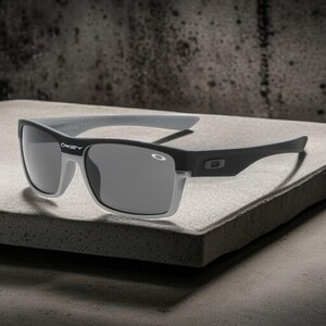 May include: Black and gray sunglasses with a rectangular frame. The sunglasses have gray lenses and gray arms. The brand name "Oakley" is visible on the frame. The sunglasses are displayed on a gray surface.