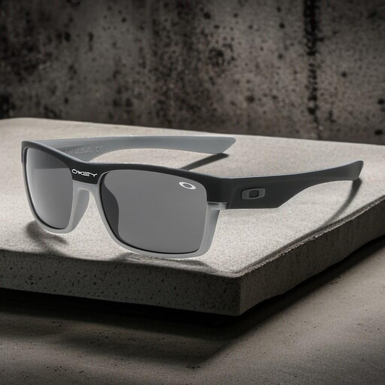 May include: Black and gray sunglasses with a rectangular frame. The sunglasses have gray lenses and gray arms. The brand name "Oakley" is visible on the frame. The sunglasses are displayed on a gray surface.
