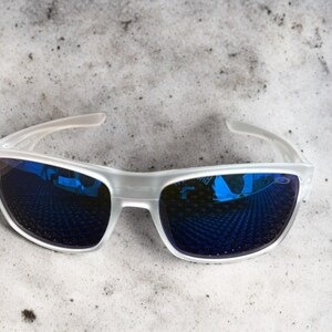May include: White-framed sunglasses with blue mirrored lenses. The sunglasses have a sleek, modern design with a semi-rimless frame. The lenses reflect a blue hue, and the arms are a translucent white.
