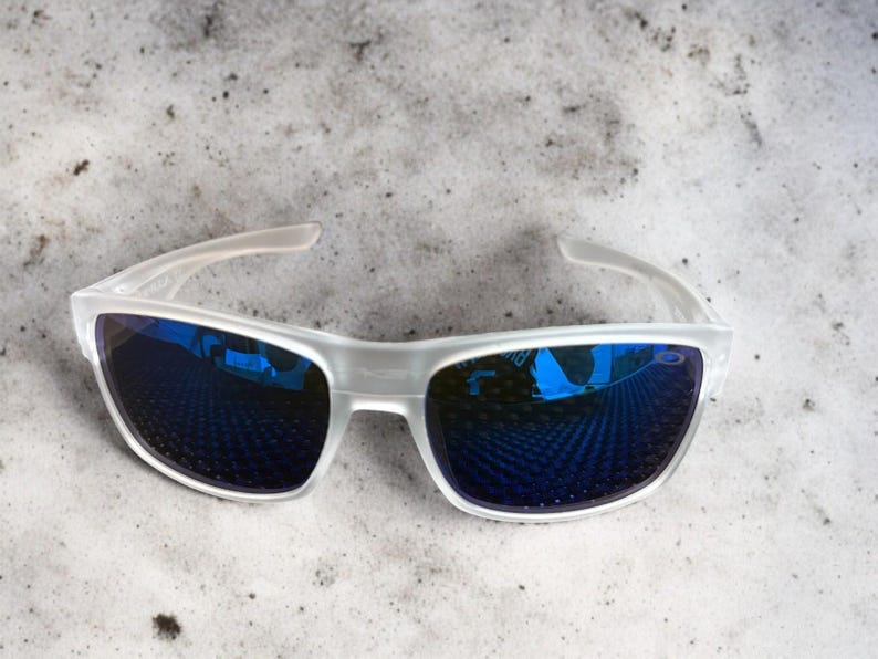 May include: White-framed sunglasses with blue mirrored lenses. The sunglasses have a sleek, modern design with a semi-rimless frame. The lenses reflect a blue hue, and the arms are a translucent white.