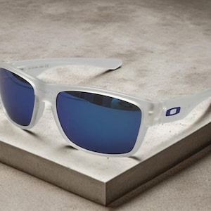 May include: White-framed sunglasses with blue mirrored lenses. The sunglasses have a rectangular shape and a small blue logo on the temple. The glasses are resting on a light gray surface.