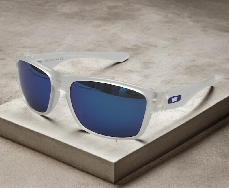 May include: White-framed sunglasses with blue mirrored lenses. The sunglasses have a rectangular shape and a small blue logo on the temple. The glasses are resting on a light gray surface.