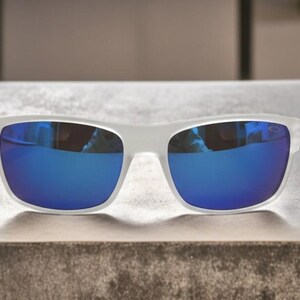 May include: White-framed sunglasses with blue mirrored lenses. The sunglasses are rectangular in shape and rest on a textured surface. The lenses reflect the surrounding environment, creating a vibrant blue hue.