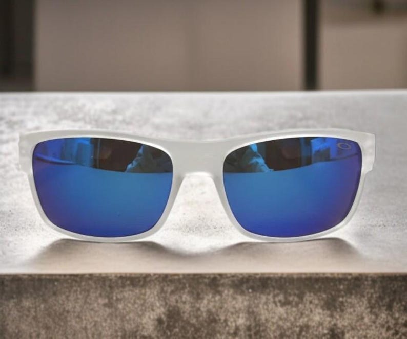May include: White-framed sunglasses with blue mirrored lenses. The sunglasses are rectangular in shape and rest on a textured surface. The lenses reflect the surrounding environment, creating a vibrant blue hue.