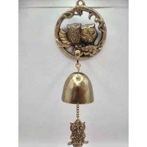 May include: A bronze-colored wind chime featuring a decorative owl design. The top has a circular frame with two owls, a bell, and a small owl pendant. The chime is suspended from a loop for hanging.