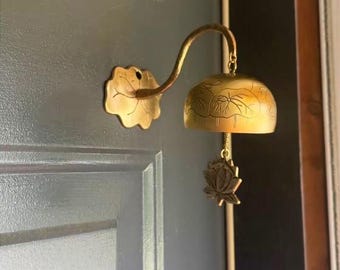 May include: A brass door chime with a lotus leaf-shaped wall mount and a curved arm supporting a bell. The bell has a floral design and a lotus flower pendant. The chime is mounted on a dark green door.