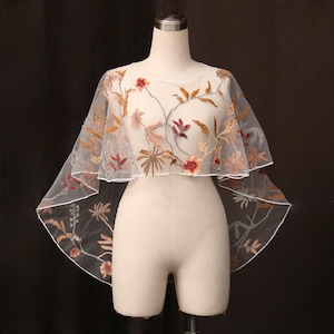 May include: A sheer white tulle capelet featuring intricate embroidery of flowers and leaves in shades of orange, brown, and red. The garment has a wide, flowing silhouette with a finished white hem, displayed on a neutral-toned mannequin.