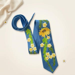 Puede incluir: Una corbata azul con bordados florales. Presenta un gran girasol amarillo y flores amarillas más pequeñas con hojas verdes. La corbata está hecha de tela lisa y se muestra sobre una superficie de color crema.