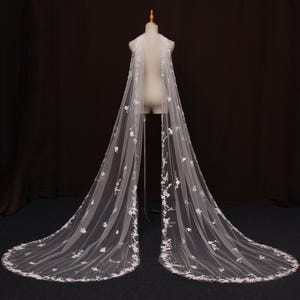 May include: A long, white bridal veil with delicate floral embroidery. The veil is displayed on a mannequin, showcasing its flowing design and intricate details. The edges are trimmed with a floral pattern.