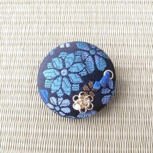 以下が含まれることがあります： 濃紺の背景に水色の花柄が織り込まれた、布製の円形ボタン。ボタンには、小さな金色の金属製フラワーチャーム、極小の白いビーズ、そして2つの小さな青い石の破片が装飾としてあしらわれています。