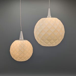 Könnte beinhalten: Zwei Pendelleuchten mit geometrisch gemusterten Lampenschirmen. Die Lampenschirme sind kugelförmig und cremefarben, mit einem Rautenmuster. Jede Leuchte hängt an einem weißen Kabel und einer weißen konischen Halterung. Der Hintergrund ist einfarbig grau.