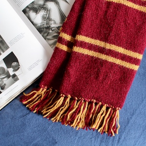 May include: A maroon knitted scarf with two yellow stripes and tassels. The scarf is lying on a blue surface.