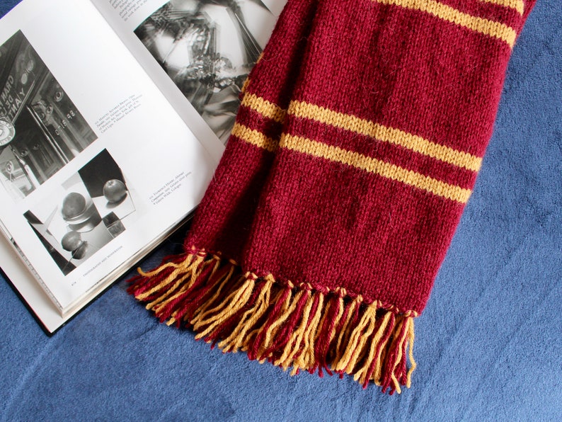 May include: A maroon knitted scarf with two yellow stripes and tassels. The scarf is lying on a blue surface.