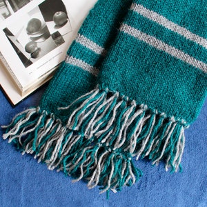 May include: A green and gray striped knitted scarf with long fringe tassels. The scarf is folded in half and the fringe is visible on both ends.