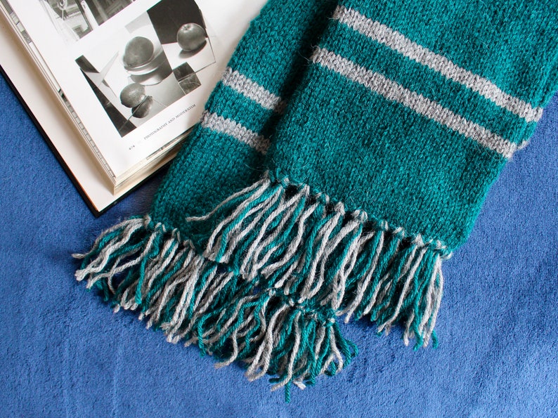 May include: A green and gray striped knitted scarf with long fringe tassels. The scarf is folded in half and the fringe is visible on both ends.