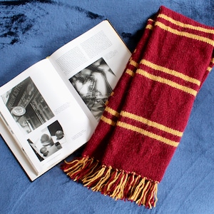 May include: A red and gold striped scarf with tassels. The scarf is lying on a blue velvet surface next to an open book.