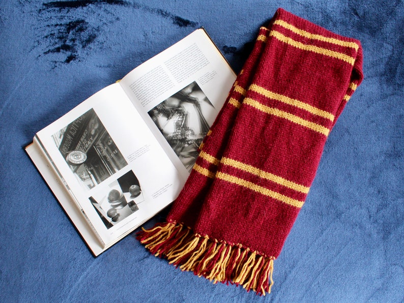 May include: A red and gold striped scarf with tassels. The scarf is lying on a blue velvet surface next to an open book.