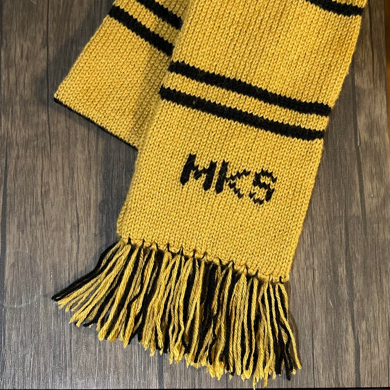 May include: A yellow and black knitted scarf with a fringe. The scarf has two black stripes and the word "H.K.S." embroidered in black.