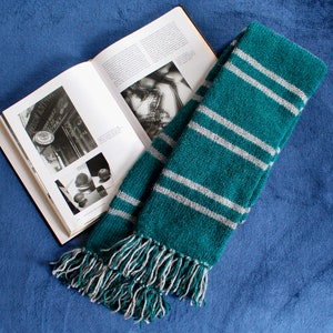 May include: A green and gray striped scarf with fringe on the ends. The scarf is folded and lying on a blue surface.