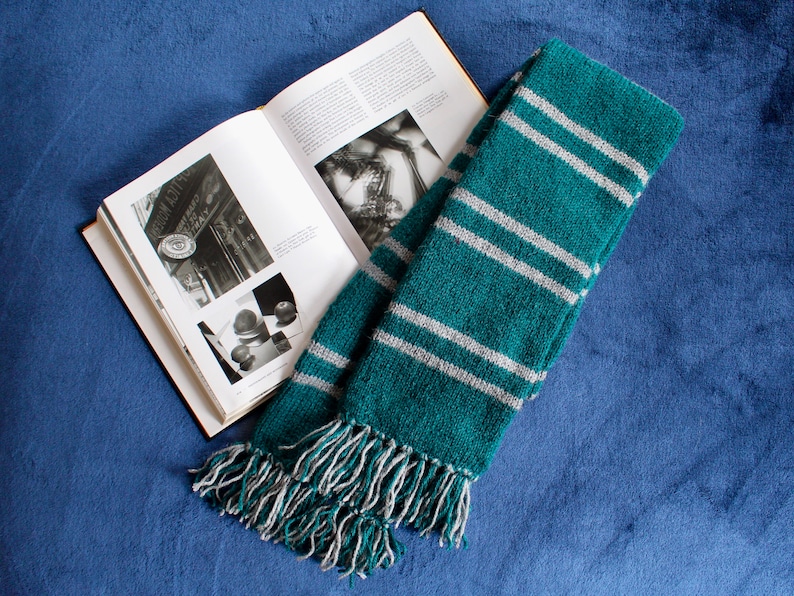 May include: A green and gray striped scarf with fringe on the ends. The scarf is folded and lying on a blue surface.