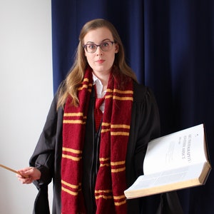 May include: A woman wearing a black robe with a red and gold striped scarf, holding a book and a wooden wand. The book has the title "STANDARD BOOK OF SPELLS" on the cover.