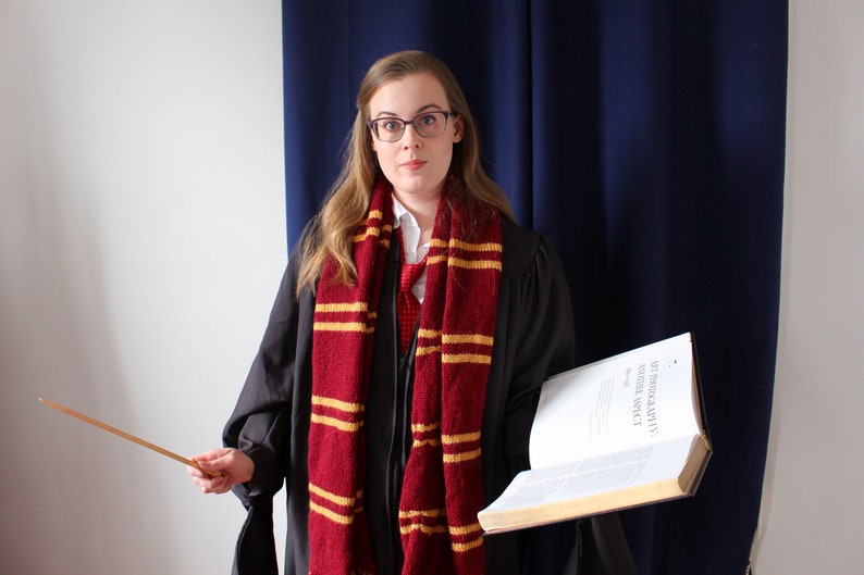 May include: A woman wearing a black robe with a red and gold striped scarf, holding a book and a wooden wand. The book has the title "STANDARD BOOK OF SPELLS" on the cover.