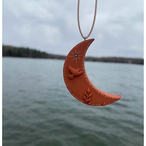 Könnte beinhalten: Ein Anhänger in Form eines halbmondförmigen Mondes in gebranntem Orange mit einer cremefarbenen Kordel. Der Anhänger zeigt ein Vogel-, Stern- und Blattdesign mit türkisfarbenem Akzent. Der Anhänger hängt vor einem See.