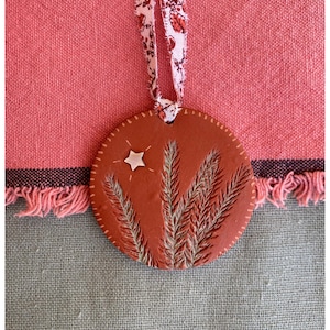 Op de afbeelding: Een rond, terracotta-kleurig ornament met een ster- en tarweontwerp, hangend aan een bloemenlint. Het ornament heeft een textuur en is rondom de rand gestikt. De achtergrond is een roze en grijze stof.