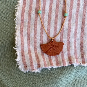 May include: A necklace featuring a brown fan-shaped pendant on a tan cord, with two turquoise beads. The necklace is displayed on a fabric with red and white stripes. The pendant is a decorative item.