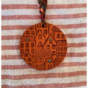 May include: A round, terracotta-colored ornament featuring a detailed relief of a village scene with houses and windows. The ornament has a decorative ribbon for hanging. The background is a striped fabric.