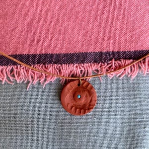May include: A circular terracotta-colored clay pendant featuring a textured, radial pattern and a small, centered turquoise bead. The pendant is suspended from a thin, light brown leather cord, creating a simple and earthy necklace design.