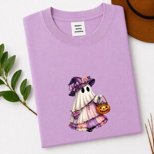 May include: A light purple t-shirt featuring a graphic of a ghost wearing a purple witch hat with a pink rose, a white and pink ruffled dress, and holding a small orange jack-o'-lantern bucket. The inner neck label reads Modern desing workshop.