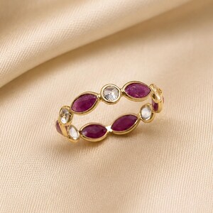 May include: A gold ring featuring alternating oval-cut ruby and round-cut diamond gemstones. The ring is set on a cream-colored satin background, showcasing the jewelry's design and color contrast. The ring's gemstones are set in a repeating pattern.