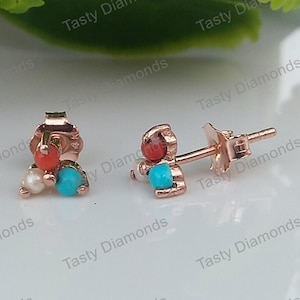 May include: Rose gold stud earrings with a cluster of colorful gemstones. Each earring features a turquoise, coral, and pearl-like stone. The earrings are displayed on a white surface.