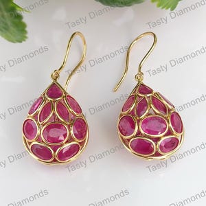 May include: A pair of gold drop earrings. Each earring has a teardrop shape, filled with oval-cut, deep red gemstones. The gemstones are set within a gold filigree design, creating a delicate and luxurious appearance.